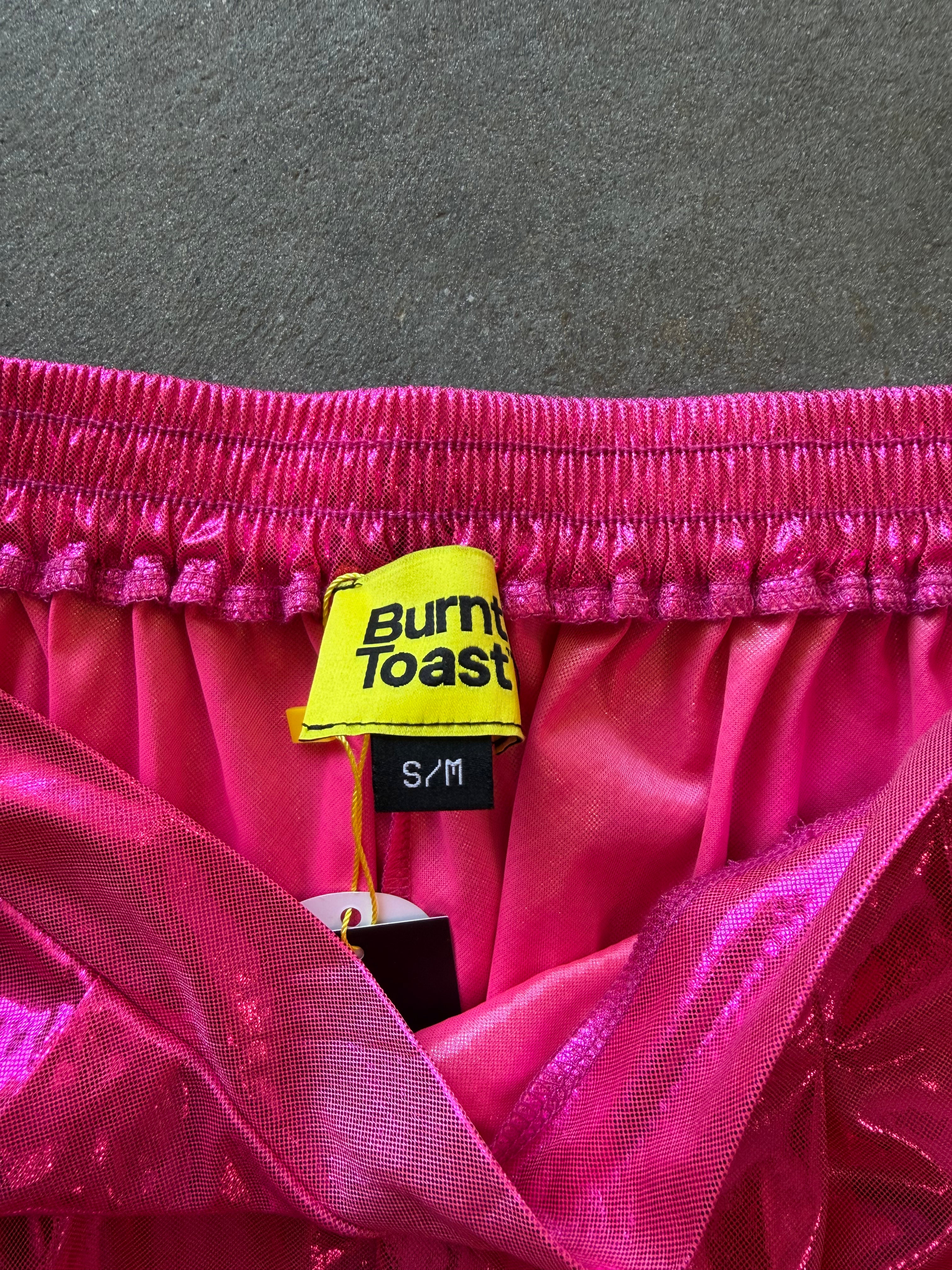 Close-up of a hot pink satin garment with a yellow Burnt Toast brand tag and black S/M size label, laid against a dark gray textured background