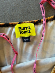 Close-up of a Burnt Toast garment showing a bright yellow brand label, a black S/M size tag, pink branded drawstring straps, and a beige fabric body on a carpeted floor