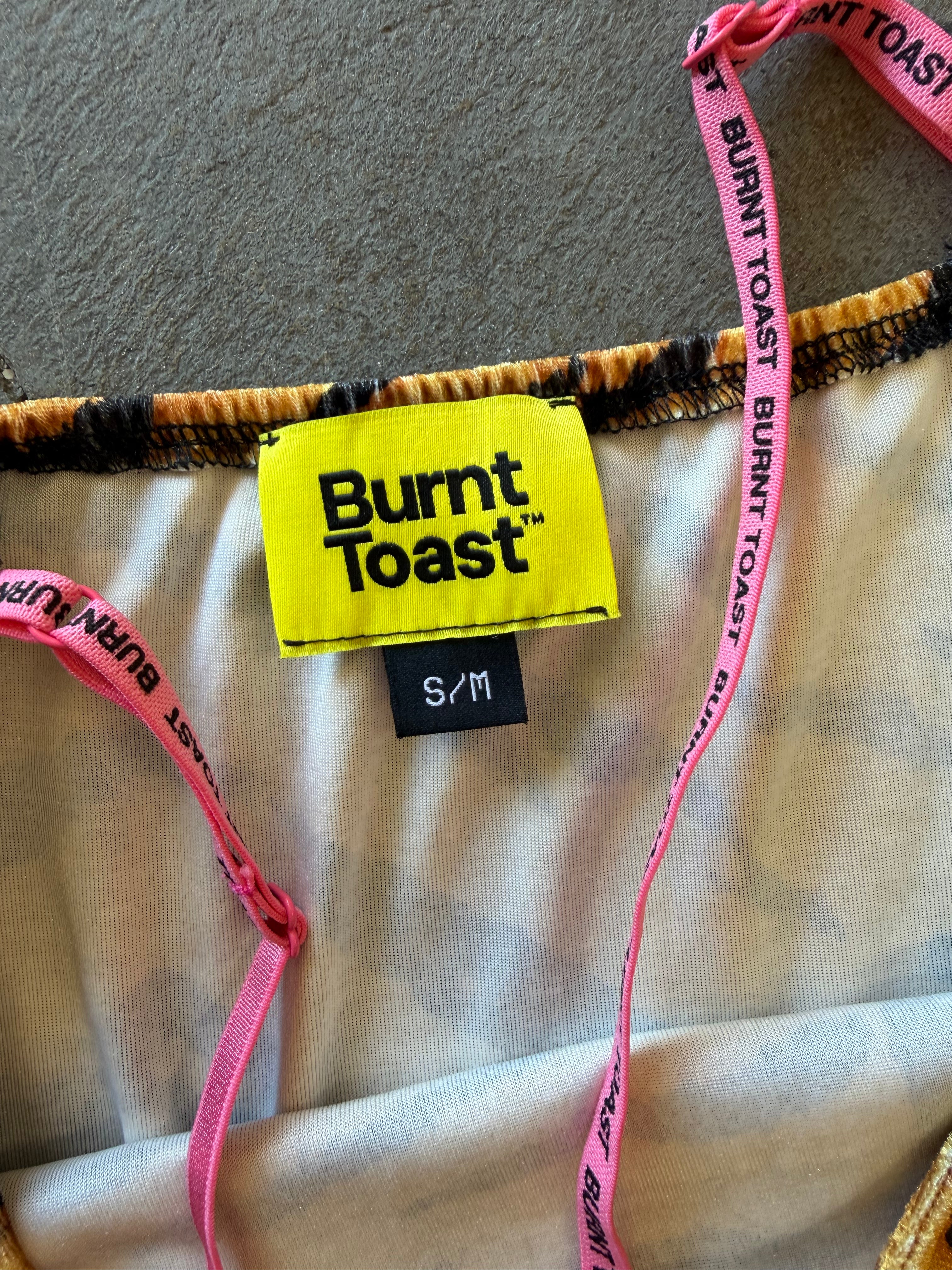 Close-up of a Burnt Toast garment showing a bright yellow brand label, a black S/M size tag, pink branded drawstring straps, and a beige fabric body on a carpeted floor