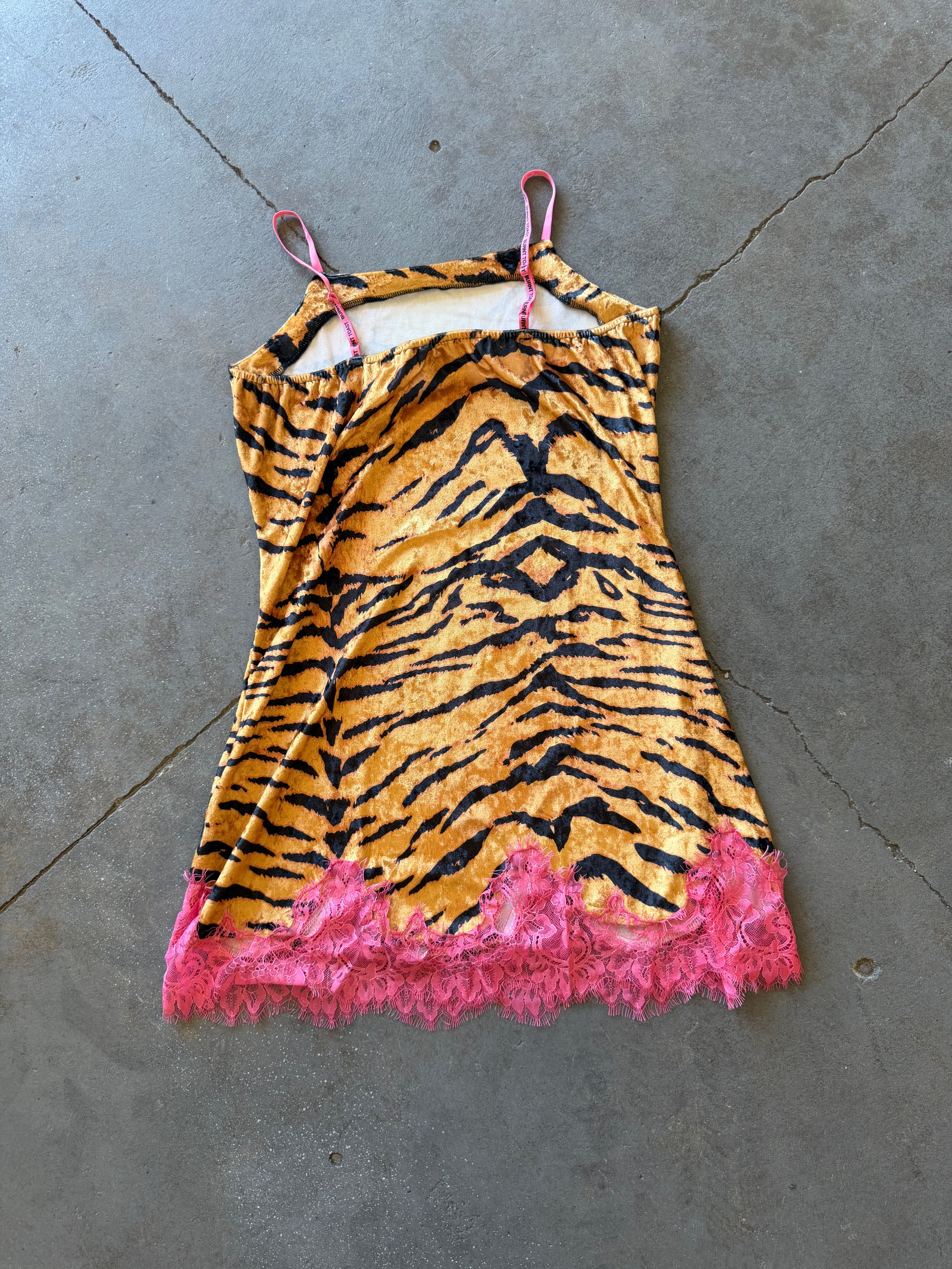 A sleeveless camisole-style dress with a tiger stripe print in orange and black, featuring bright pink lace trim along the hem and matching pink straps, laid flat on a gray concrete floor