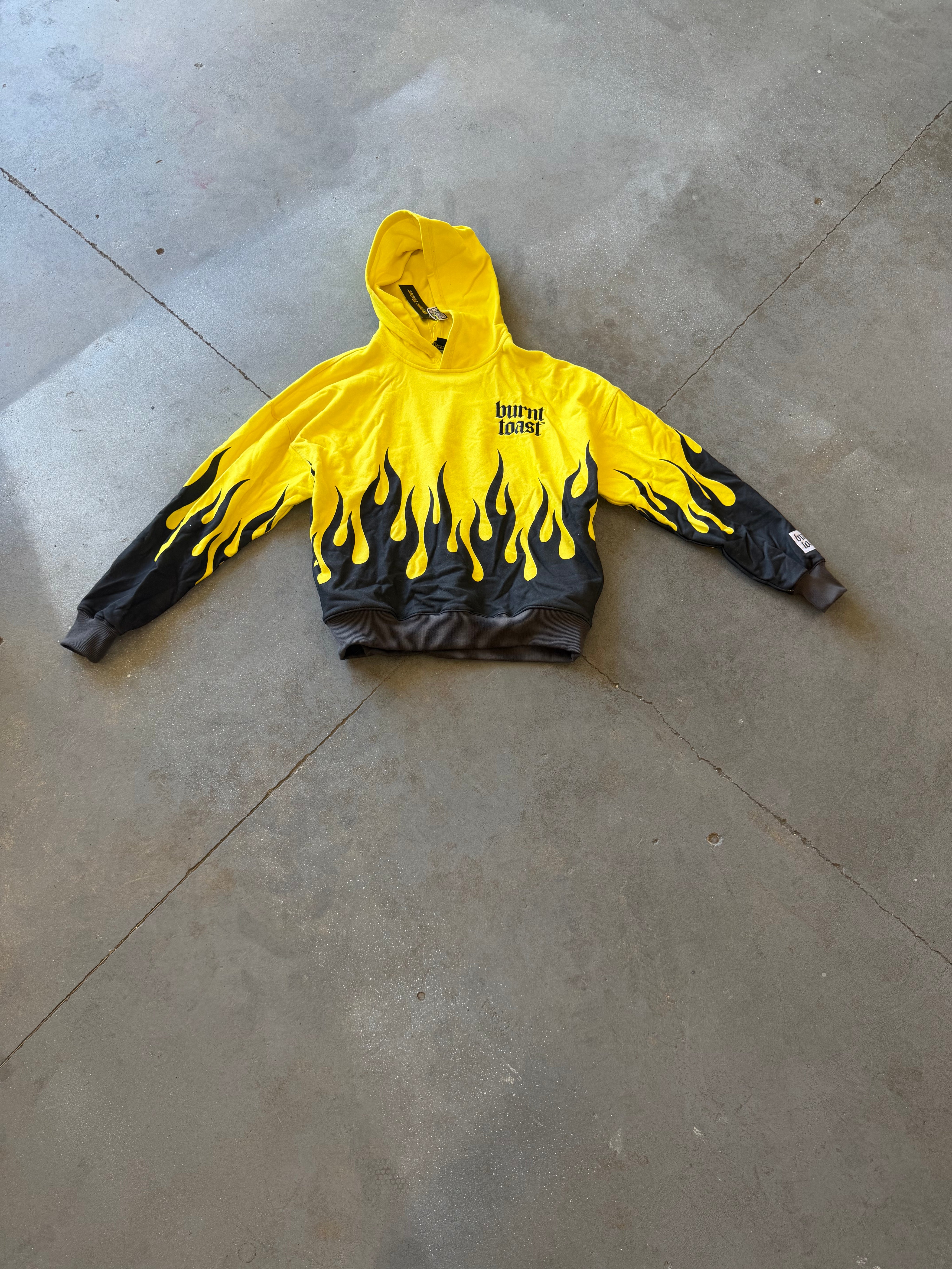 A yellow and black flame-print hoodie with 'Burnt Toast' printed on the chest, laid flat on a gray concrete floor