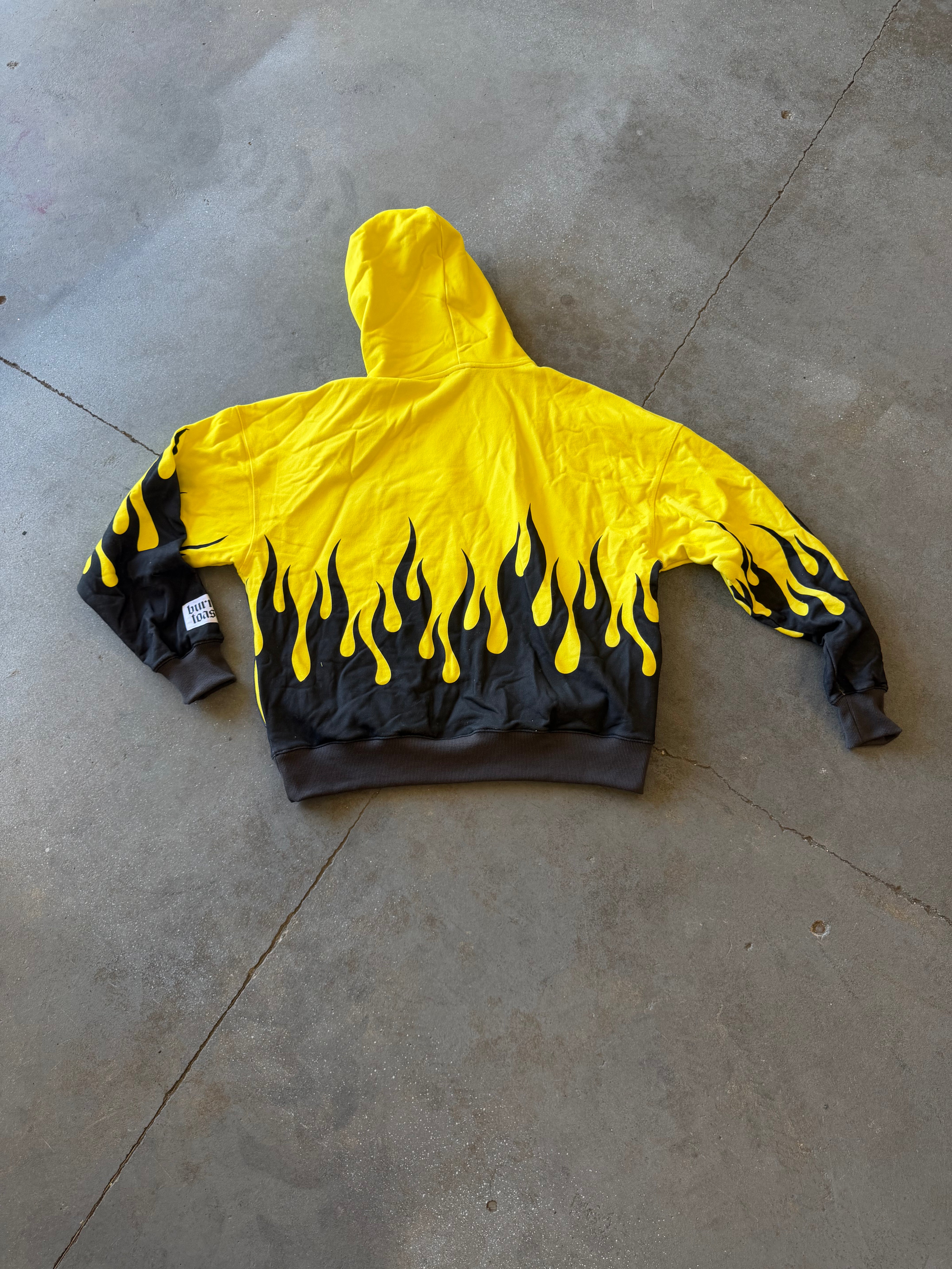 A yellow hooded sweatshirt with a black flame print along the bottom hem and cuffs, laid flat on a gray concrete floor