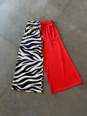 A pair of wide-leg cropped pants laid flat on a gray concrete floor, featuring a split design with a black and white zebra print on one leg and solid bright red on the other
