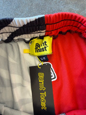Close-up of the waistband of Burnt Toast shorts, showing a color-blocked pattern of red, black, white, and gray ribbed fabric, with a yellow and black brand tag and a black hangtag