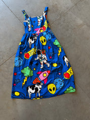 A blue sleeveless dress with a colorful space-themed print featuring cartoon astronauts, rockets, aliens, cows, clouds, and stars, laid flat on a gray concrete floor