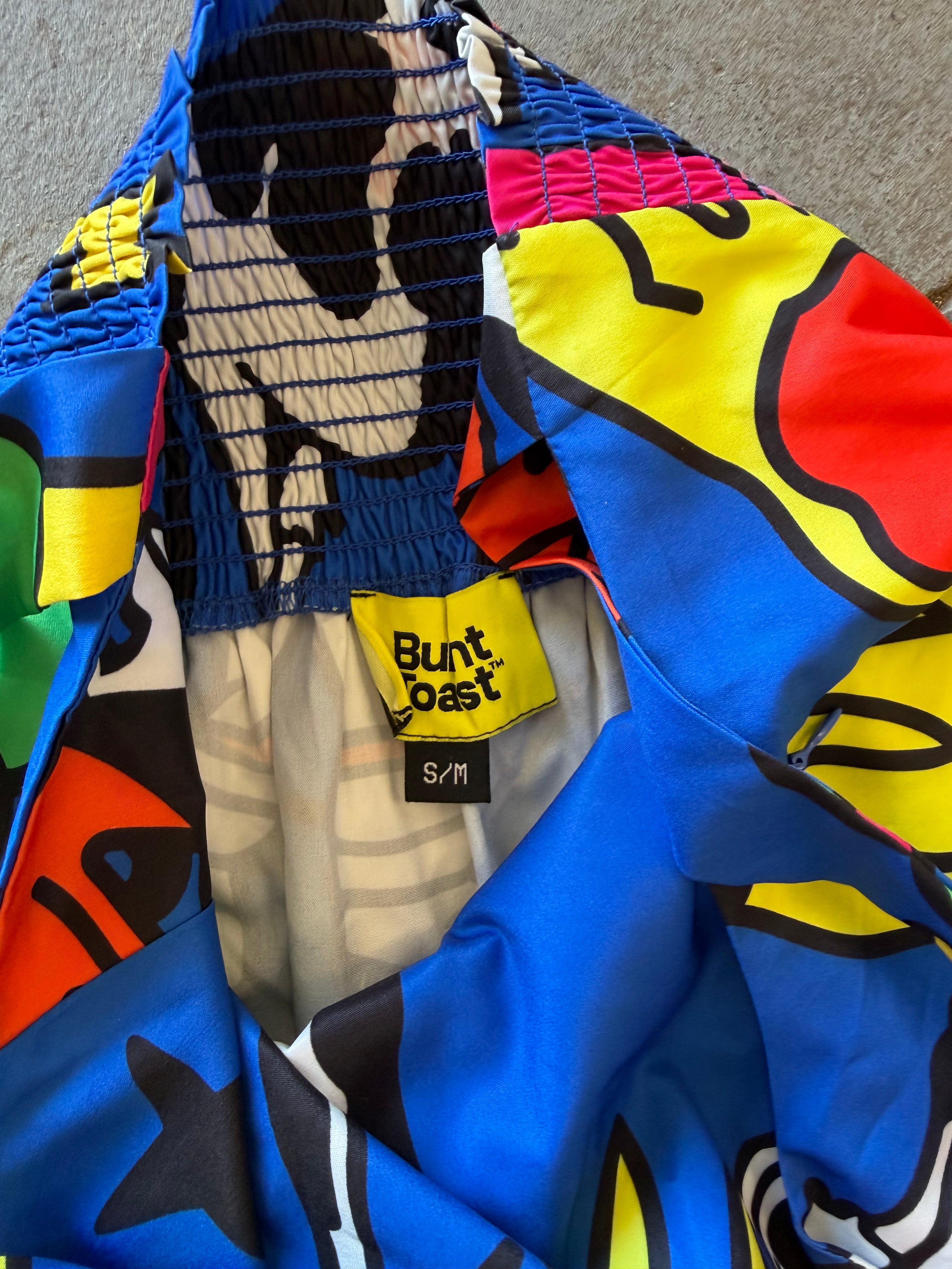 Close-up of a colorful Bunt-Toast garment, likely a jacket or coat, featuring a vibrant abstract print in blue, yellow, red, black, and white. The elasticized collar is open, revealing a black-and-white patterned lining, and a yellow Bunt-Toast brand label is visible