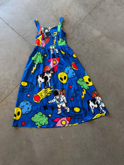 A sleeveless blue sundress with a colorful cartoon space print, featuring astronauts, aliens, rockets, and cows, laid flat on a concrete floor