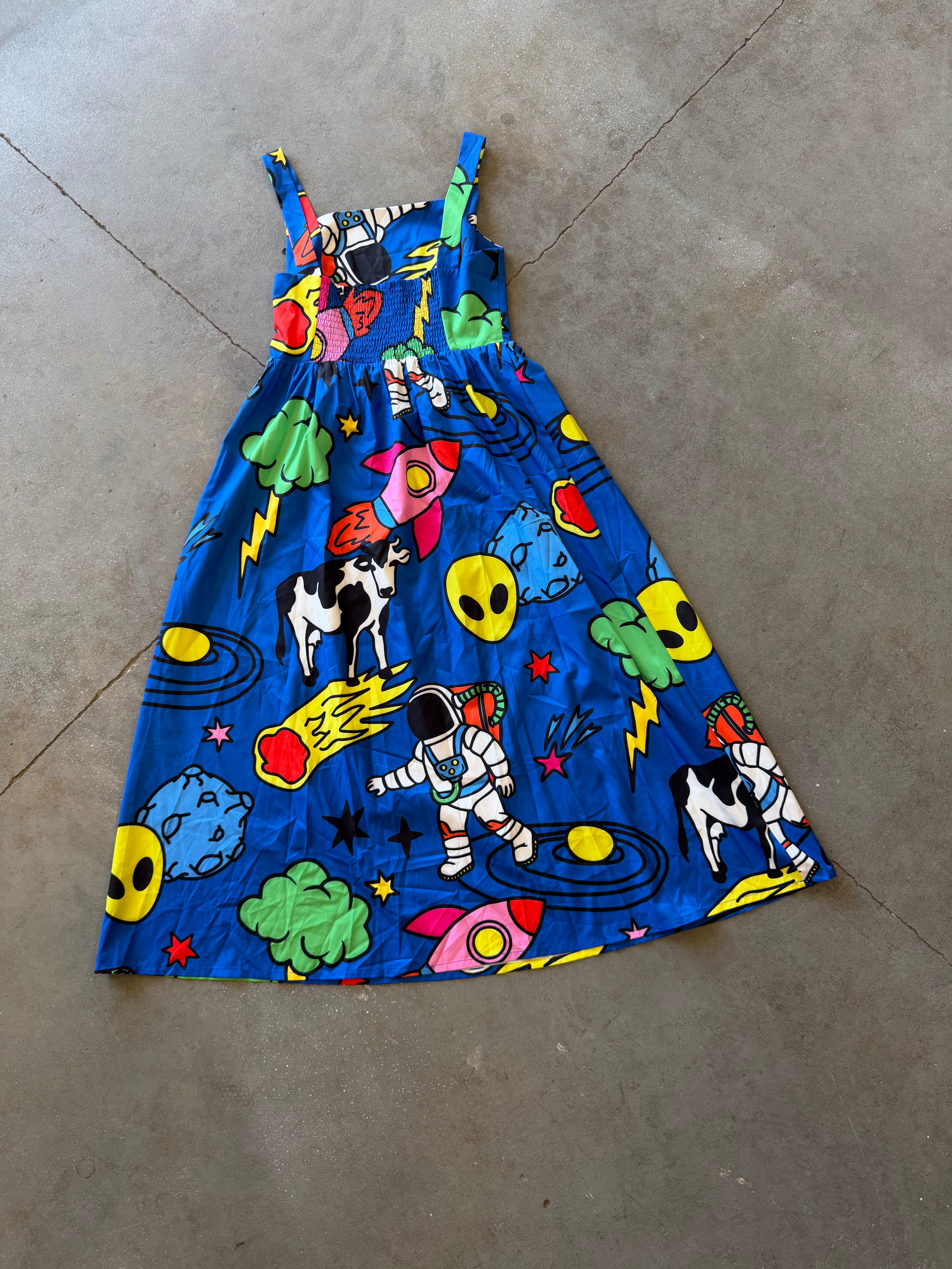 A sleeveless blue sundress with a colorful cartoon space print, featuring astronauts, aliens, rockets, and cows, laid flat on a concrete floor