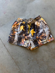 A pair of camouflage athletic shorts with a yellow drawstring and two yellow star-shaped appliqués, laid flat on a gray concrete floor