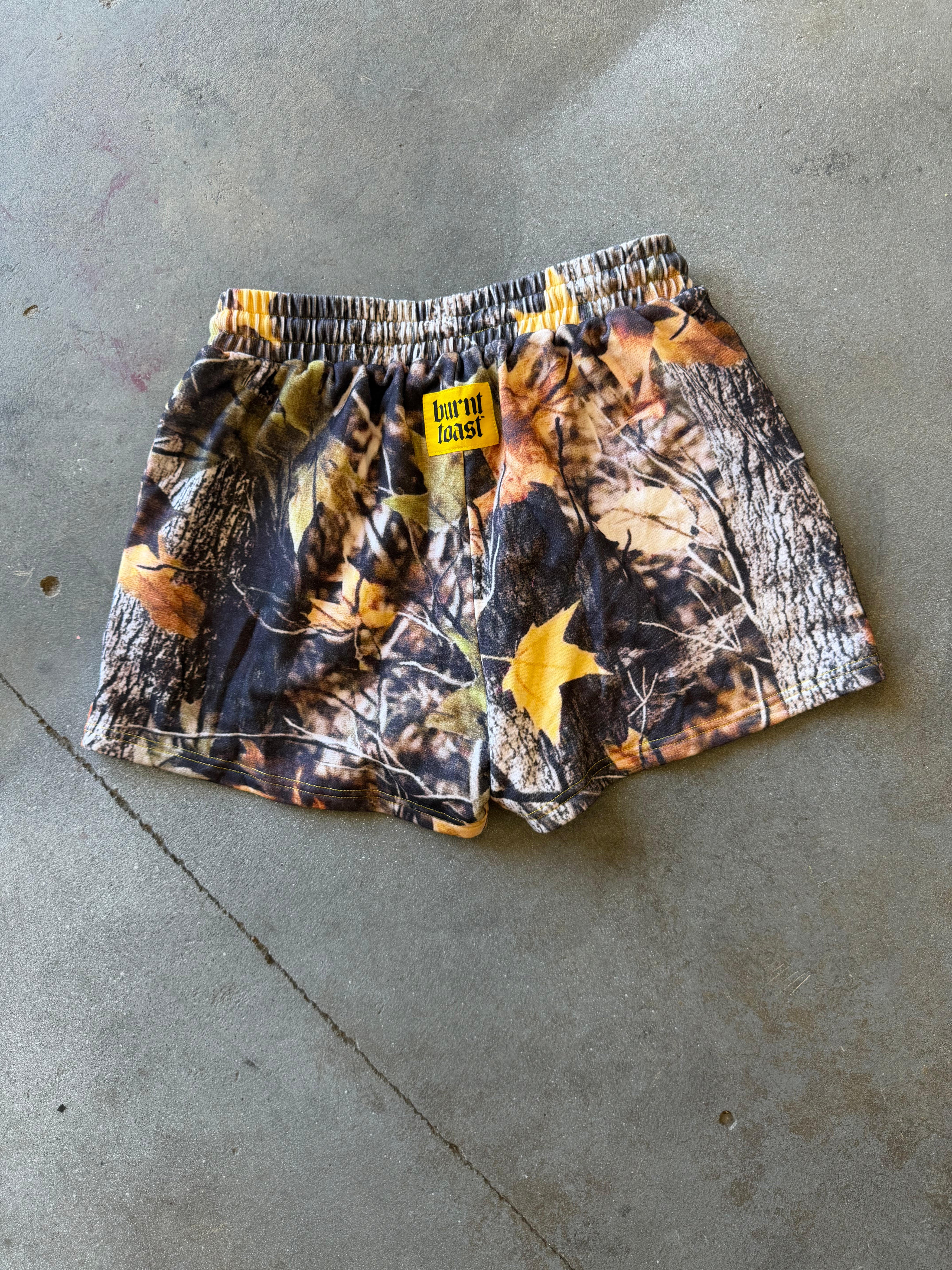 A pair of camouflage shorts with a yellow 'Bourne Boots' brand patch, laid flat on a gray concrete floor