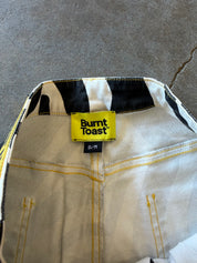 Close-up of the inside waistband of a pair of jeans, showing a yellow Burnt Toast brand label, black and white striped inner waistband, and off-white denim fabric with yellow stitching on a gray concrete floor