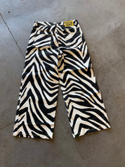 Zebra-print cropped pants with a tie waist, laid flat on a gray concrete floor. The fabric features a bold black and cream zebra pattern, and a small yellow brand label is visible at the waistband