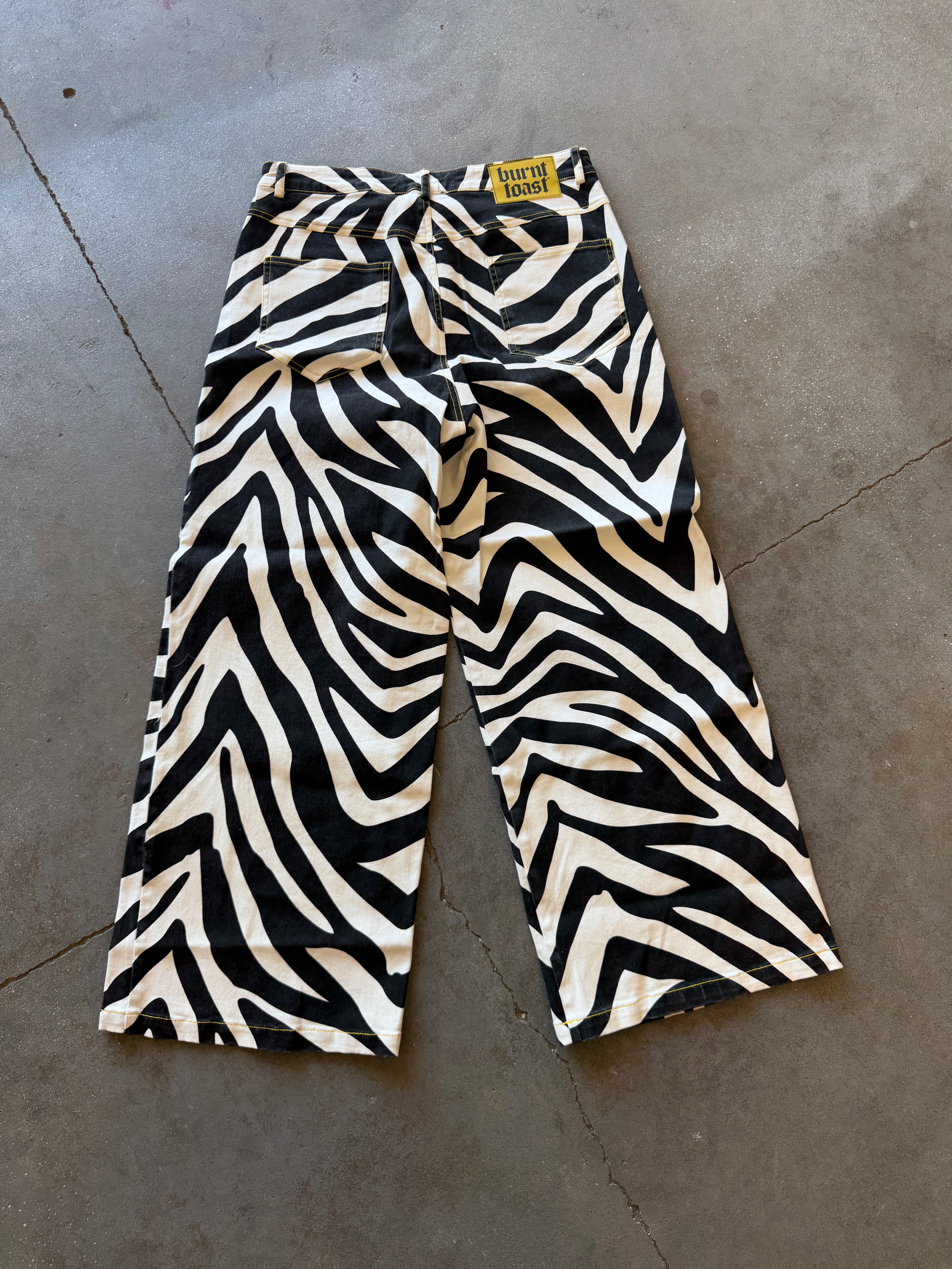 Zebra-print cropped pants with a tie waist, laid flat on a gray concrete floor. The fabric features a bold black and cream zebra pattern, and a small yellow brand label is visible at the waistband