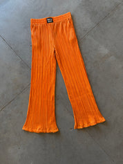 Bright orange pleated wide-leg pants laid flat on a gray concrete floor. The pants feature a textured vertical pleat pattern and a black brand label at the waistband