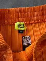 Close-up of the inside waistband of an orange pleated pants by Burnt Toast, showing the yellow brand label, black size tag, and hangtag against a dark gray fabric background