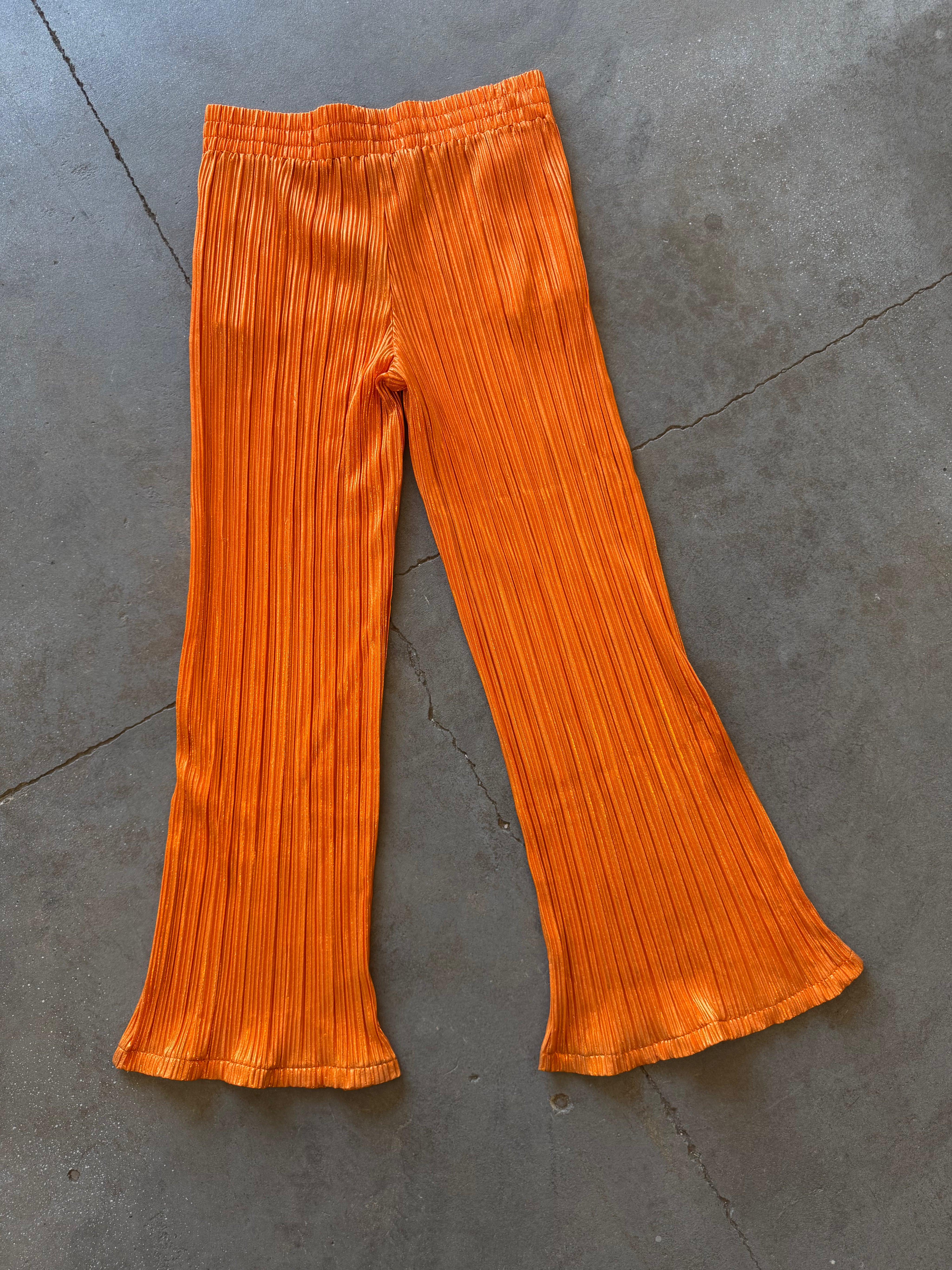 Bright orange pleated flared pants laid flat on a gray concrete floor