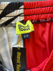 Close-up of a pair of ribbed pants showing the waistband with three color panels: black and white on the left, red and white in the middle, and solid red on the right. A neon yellow 'Burnt Toast' brand label and a black size tag reading 'S/M' are attached