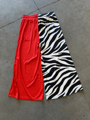 A pair of wide-leg cropped pants laid flat on a gray concrete floor, featuring a split design with one leg in solid bright red and the other leg in a black and white zebra print