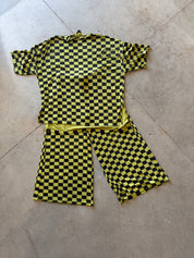 A two-piece matching set consisting of a short-sleeve top and pants, both featuring a bold yellow and black checkerboard pattern, laid flat on a concrete floor
