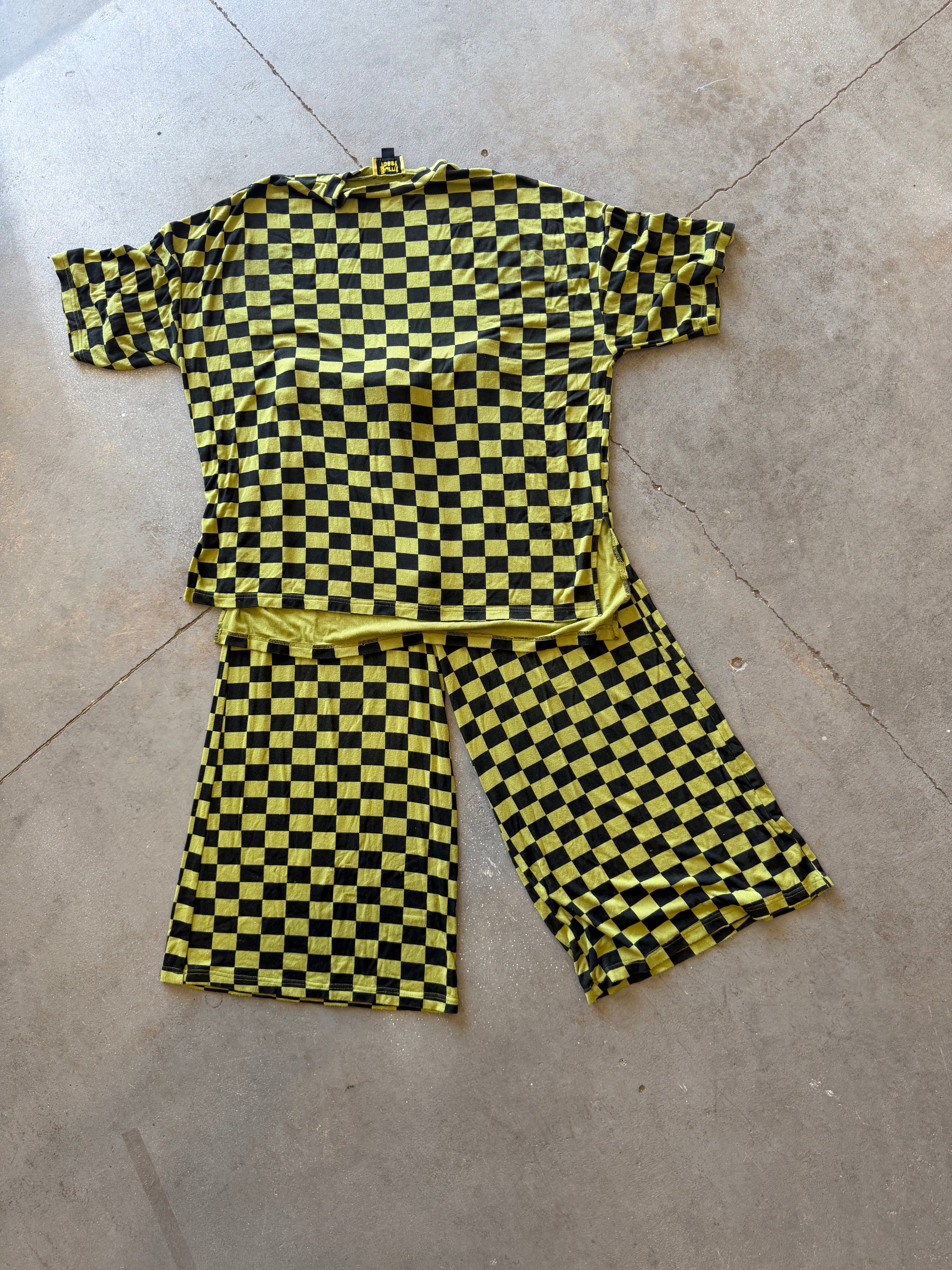 A two-piece matching set consisting of a short-sleeve top and pants, both featuring a bold yellow and black checkerboard pattern, laid flat on a concrete floor