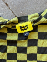 Close-up of a yellow and black checkered garment with a visible Burnt Toast brand label and size S/M, laid flat on a gray concrete surface