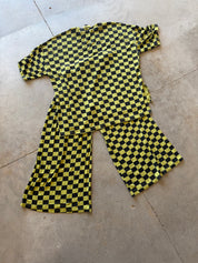 A two-piece matching set consisting of a short-sleeve top and pants, both featuring a bold yellow and black checkerboard pattern, laid flat on a concrete floor