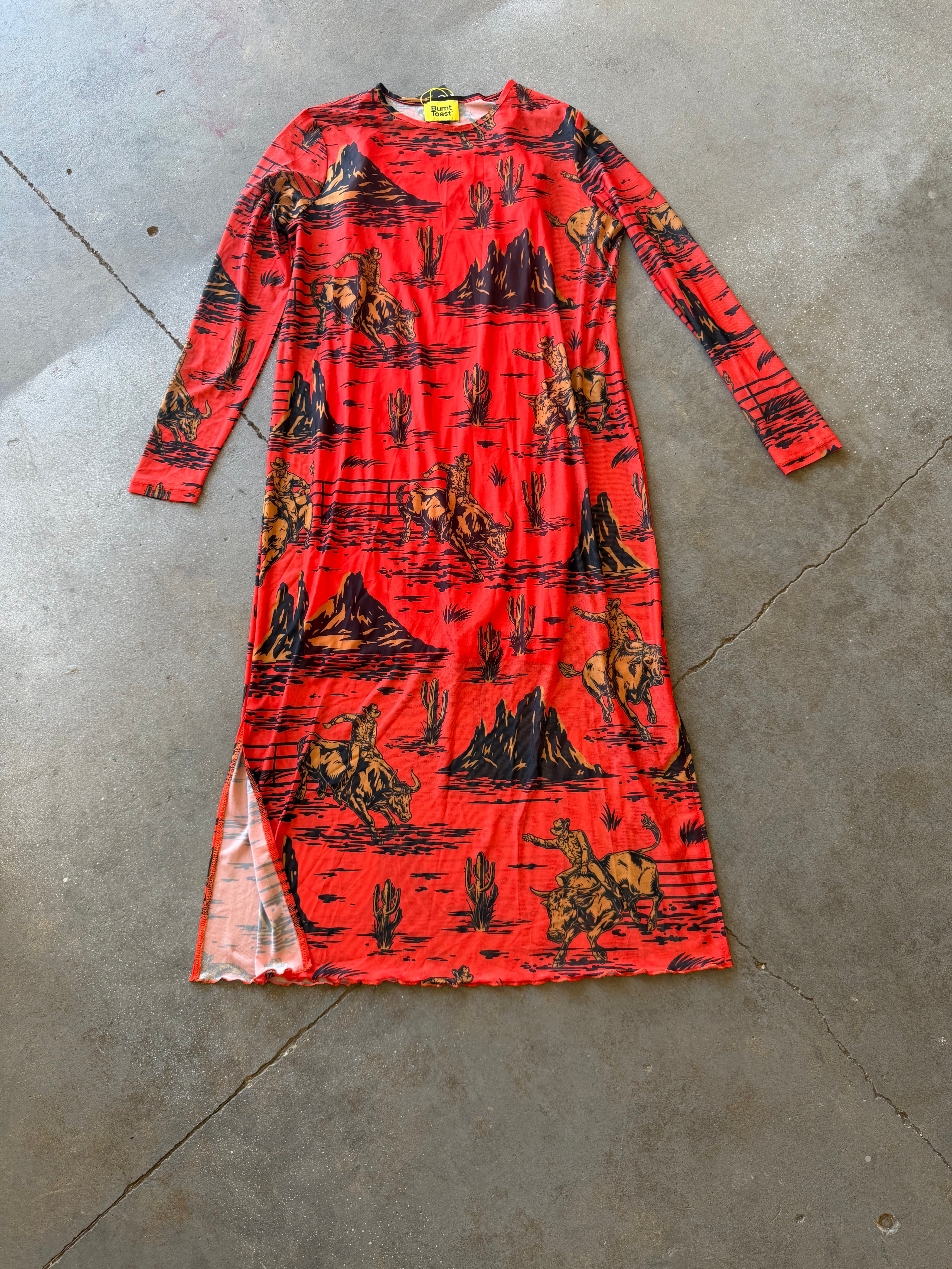 A long-sleeve red dress with a black and orange western-style print of mountains, cacti, and riders, laid flat on a gray concrete floor