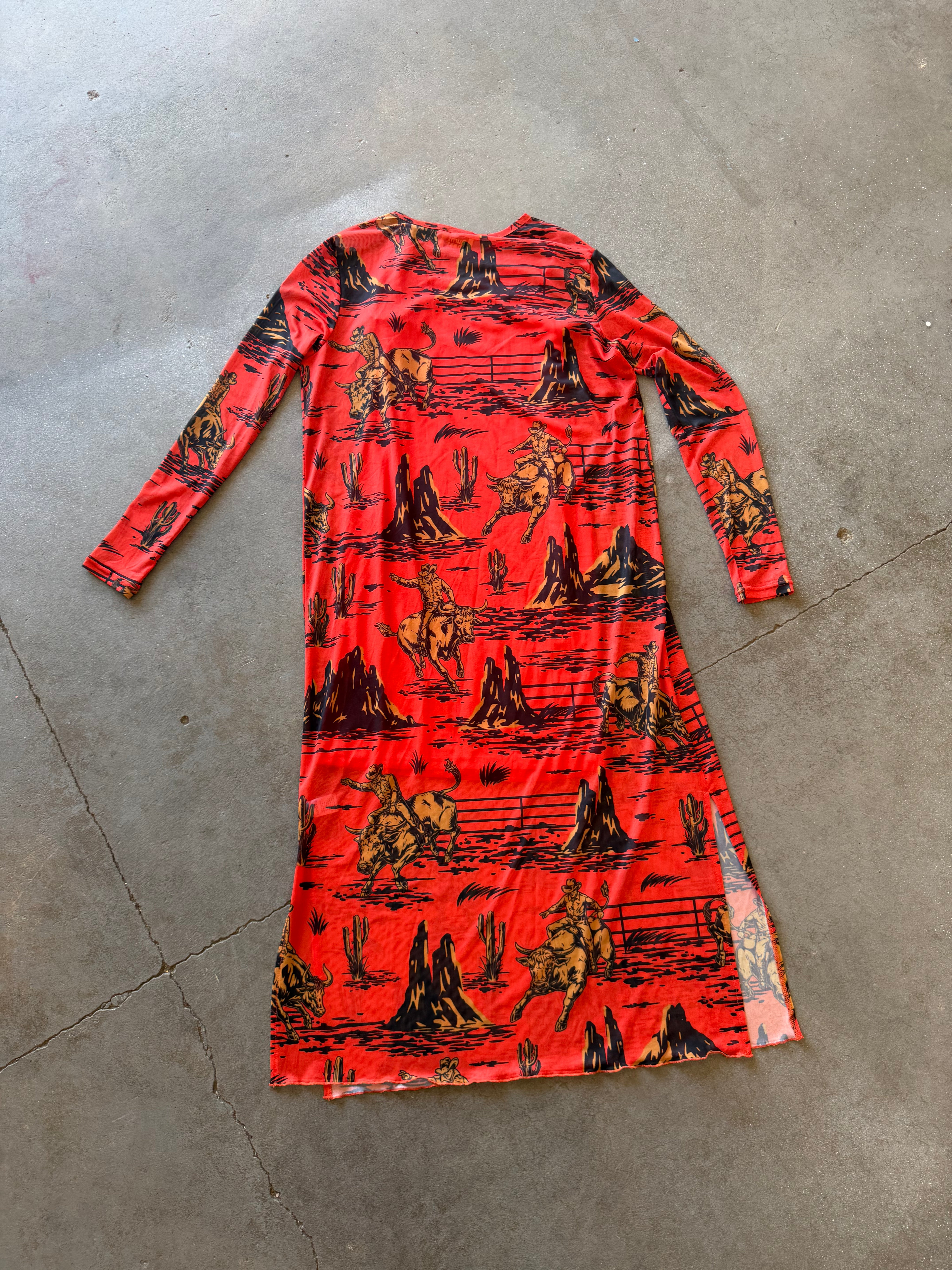A long-sleeve red dress with a black and yellow western-themed print of cowboys, horses, and mountains, laid flat on a gray concrete floor