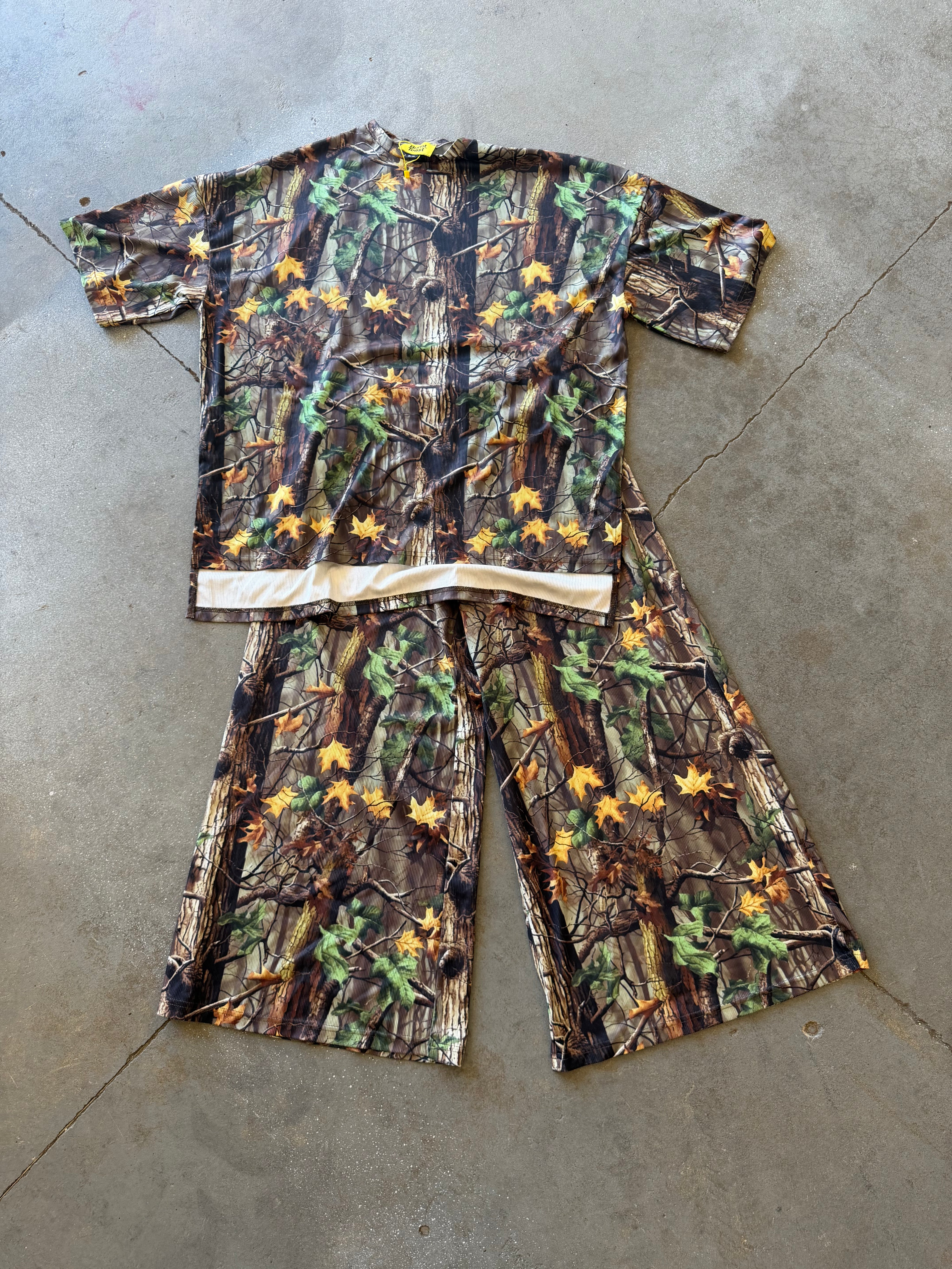 A two-piece camouflage set consisting of a short-sleeve top and matching pants, laid flat on a gray concrete floor. The camouflage pattern features brown tree bark, green leaves, and small yellow flowers, with a light beige waistband on the top
