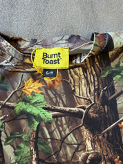 Close-up of a Burnt Toast camouflage shirt with a bright yellow brand label and size S/M, laid flat on a gray textured surface