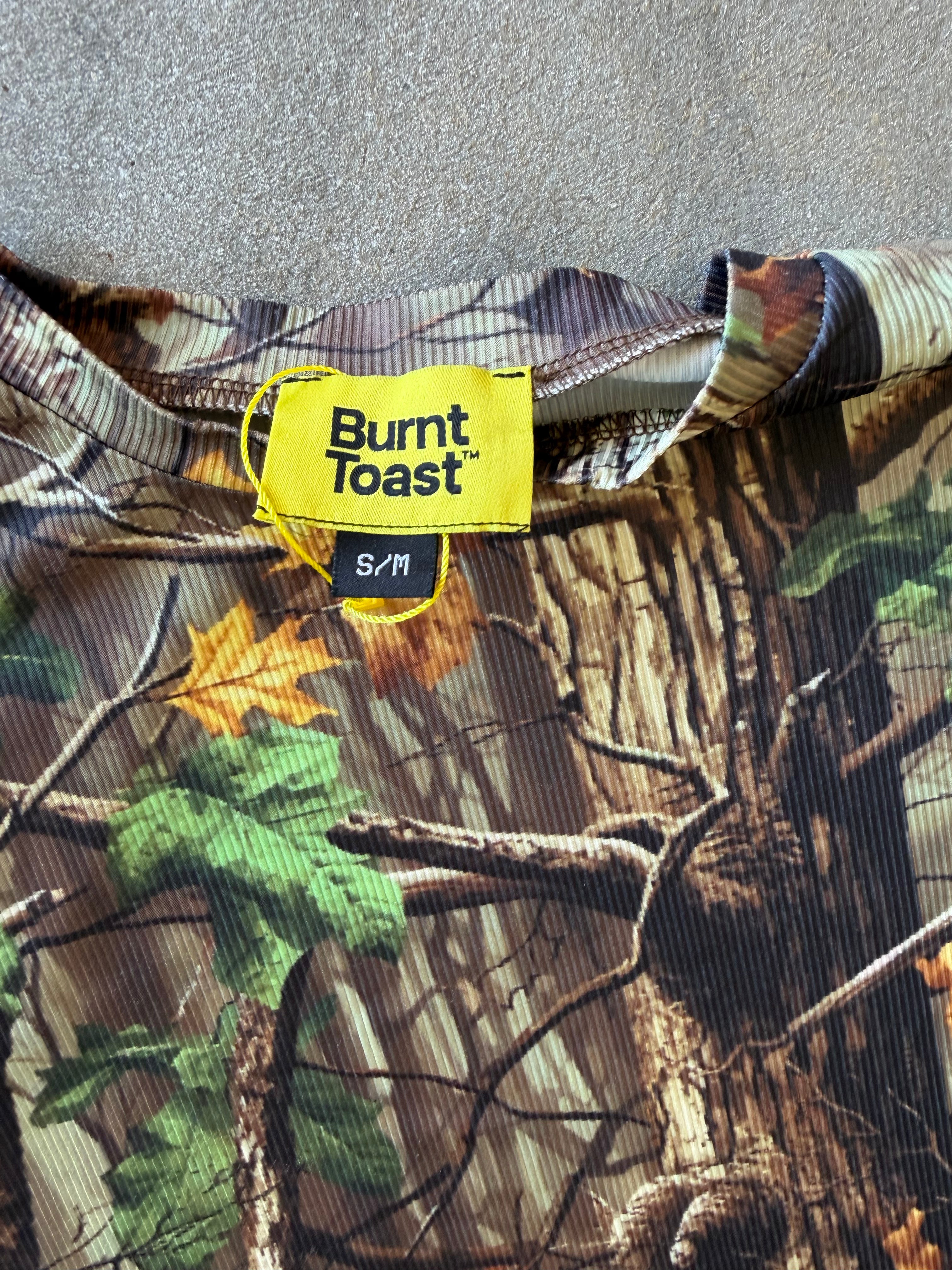 Close-up of a Burnt Toast camouflage shirt with a bright yellow brand label and size S/M, laid flat on a gray textured surface