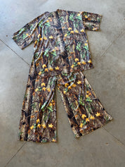 A two-piece short-sleeve pajama set laid flat on a concrete floor, featuring a woodland camouflage print with green leaves and yellow/orange autumn leaves over brown and gray tree bark