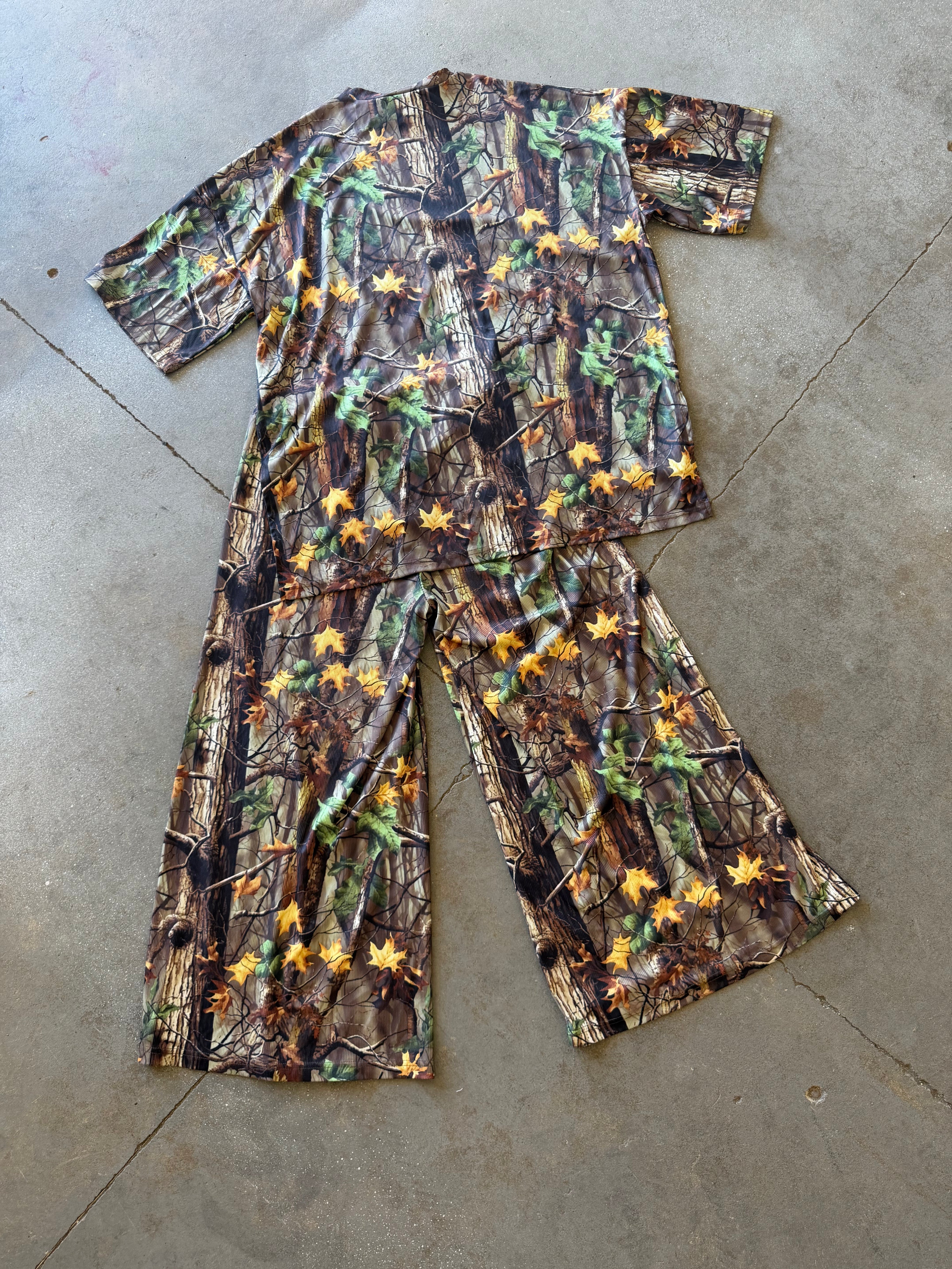 A two-piece short-sleeve pajama set laid flat on a concrete floor, featuring a woodland camouflage print with green leaves and yellow/orange autumn leaves over brown and gray tree bark