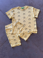 A two-piece baby biker set consisting of a short-sleeve top and matching shorts in a pale yellow color, both printed with a black smiley face pattern, laid flat on a dark gray concrete floor