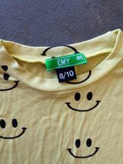 A close-up of a pale yellow ribbed knit top with a black smiley face pattern, featuring a green and black 'EMY AVE' neck label