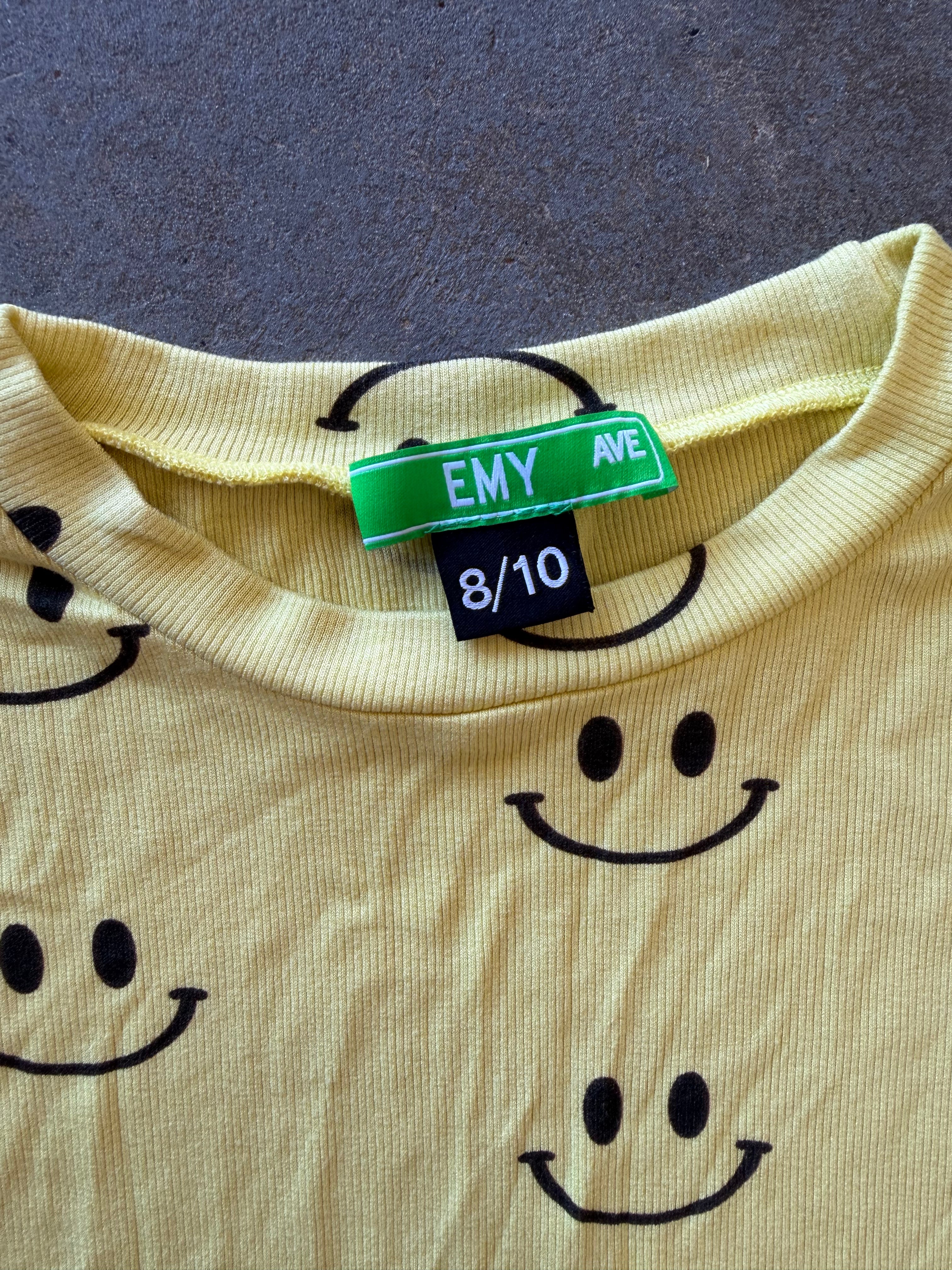 A close-up of a pale yellow ribbed knit top with a black smiley face pattern, featuring a green and black 'EMY AVE' neck label