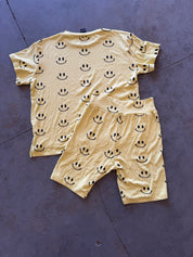 A two-piece pajama set consisting of a short-sleeve t-shirt and matching shorts, both in a pale yellow color with a black smiley face pattern, laid flat on a dark gray concrete floor