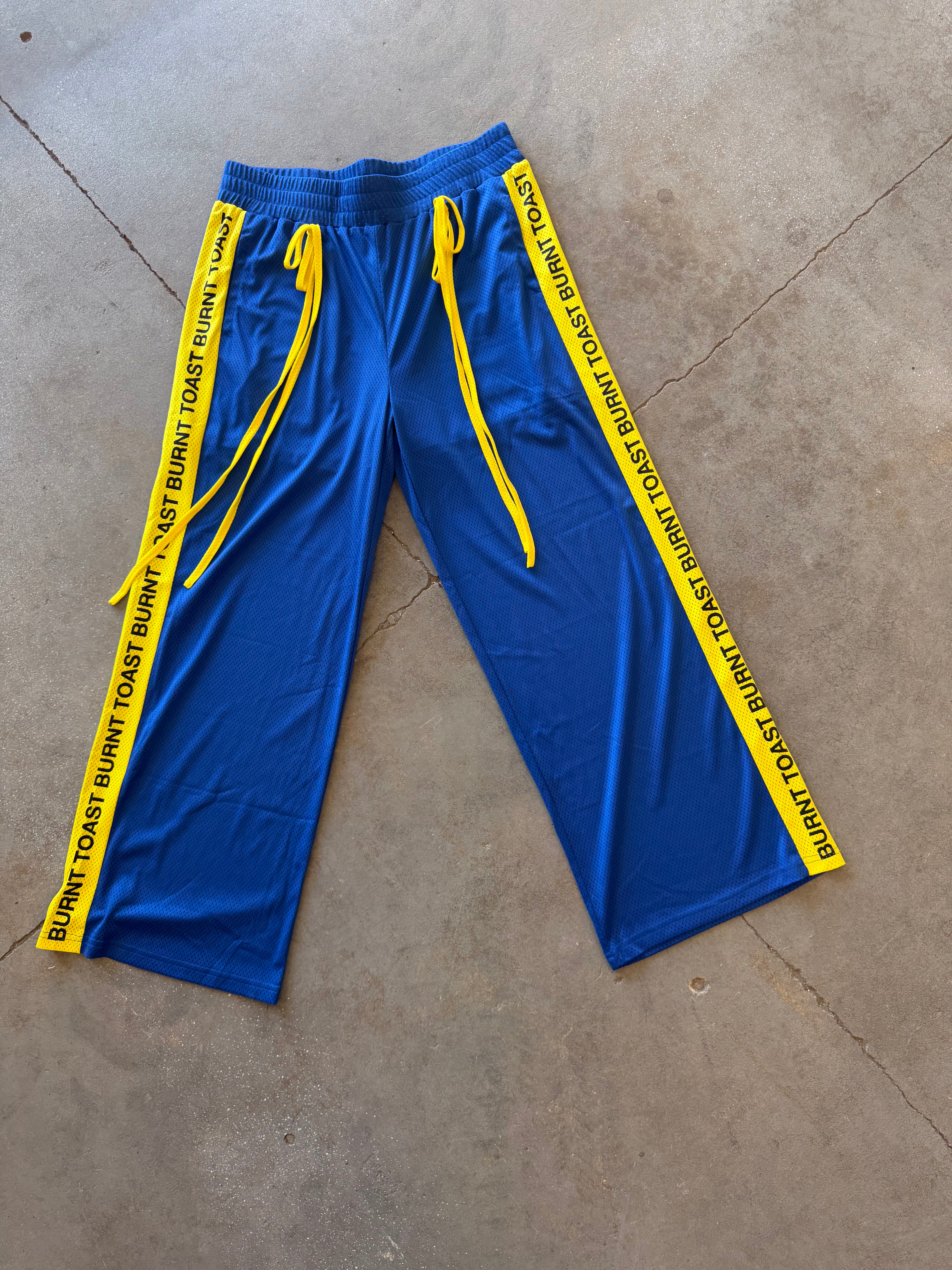 Blue athletic pants with yellow drawstrings and yellow side tape printed with the text 'BURNT TOAST' in black, laid flat on a concrete floor