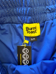 Close-up of the inner waistband of a blue athletic garment, showing a yellow Burnt Toast brand label, a black size tag reading S/M, and a black hangtag with smiley faces