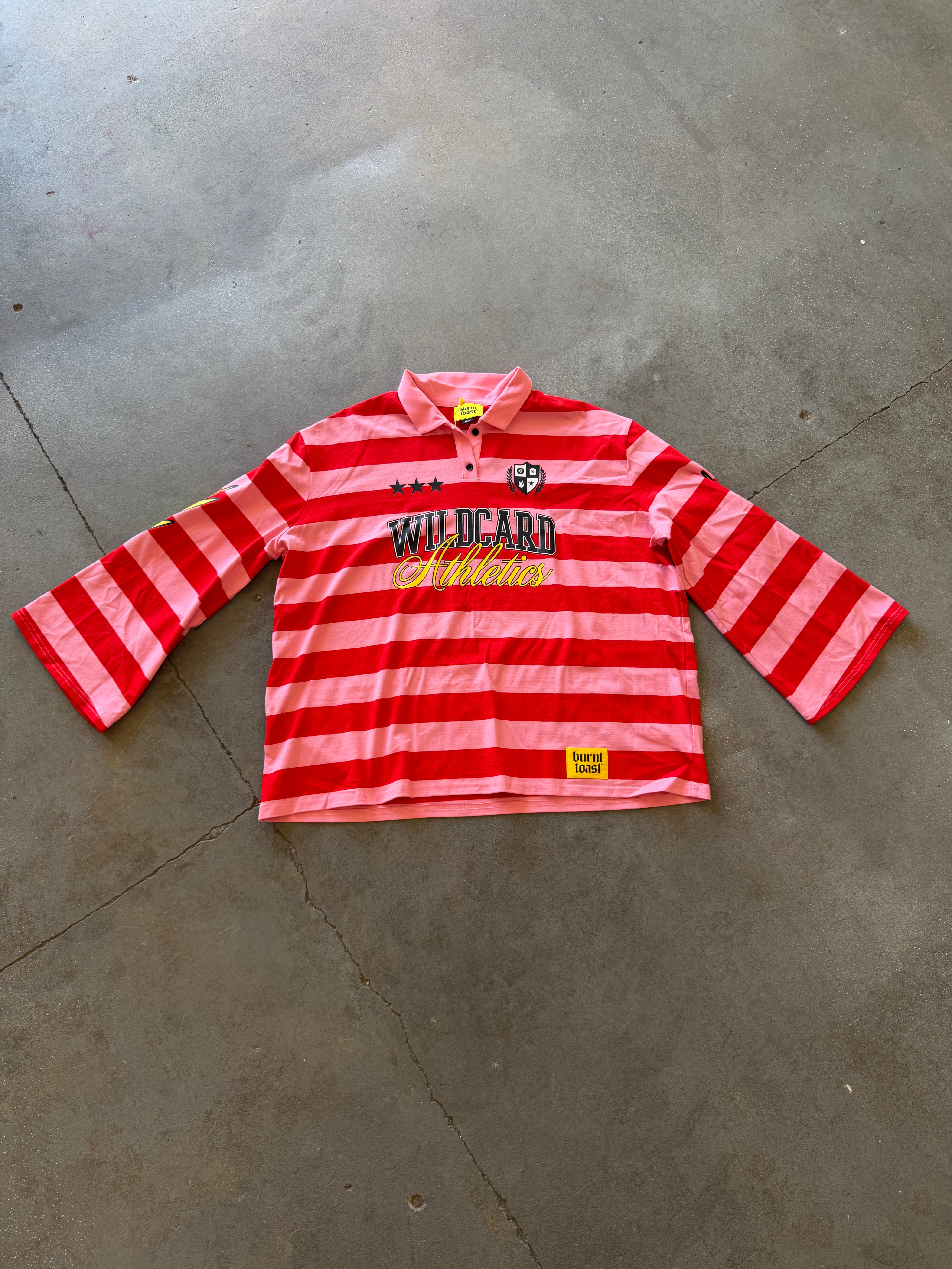 A red and pink striped long-sleeve rugby shirt with 'WILDCARD Athletics' printed on the chest, laid flat on a gray concrete floor