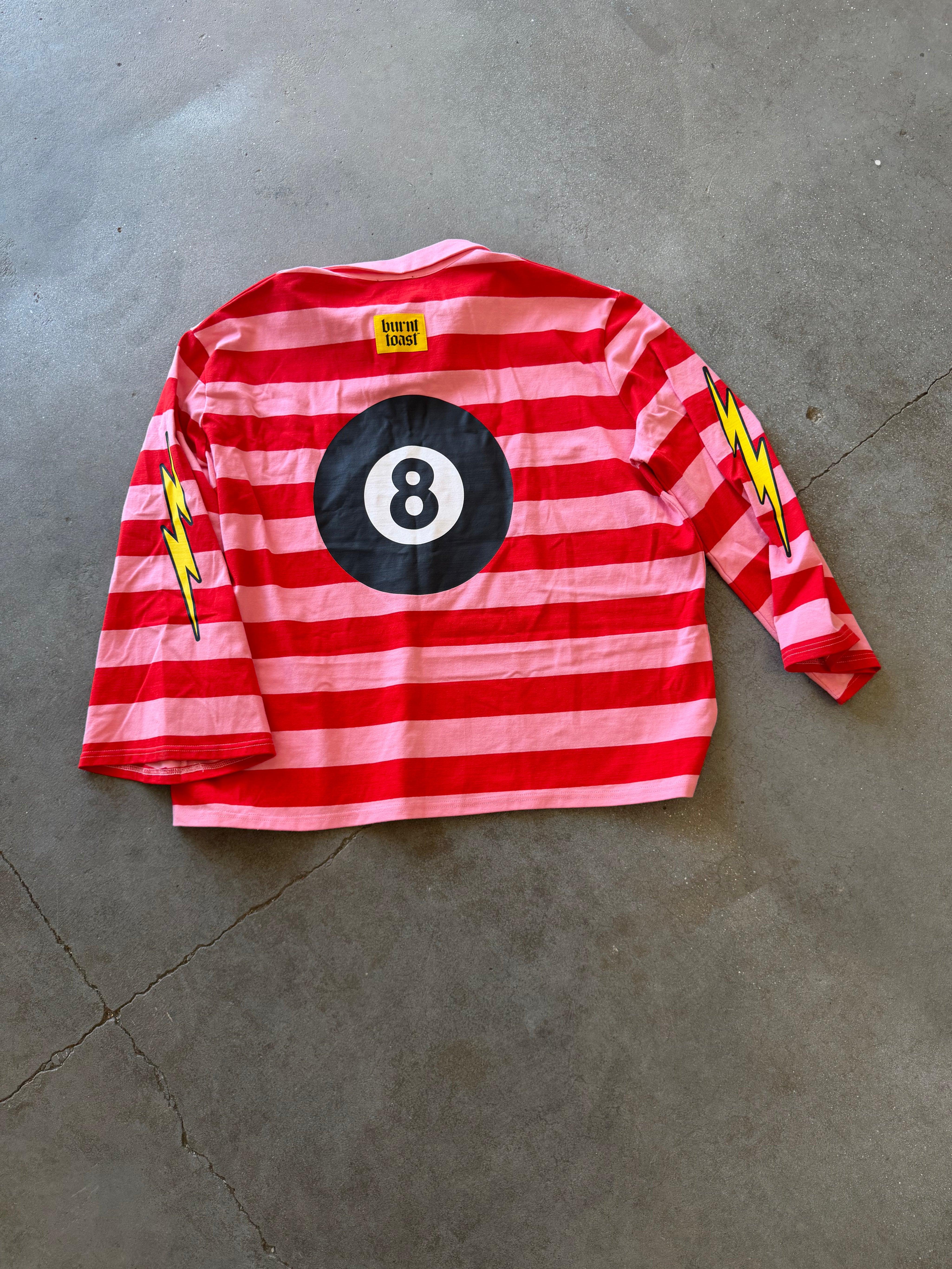 A red and pink striped long-sleeve shirt laid flat on a gray concrete floor, featuring a large black and white billiard 8-ball graphic on the back, yellow lightning bolt patches on both sleeves, and a small yellow brand label at the neck