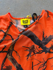 An orange short-sleeve top with a black and brown tree branch and leaf camouflage pattern, featuring a yellow Burnt Toast brand label at the collar