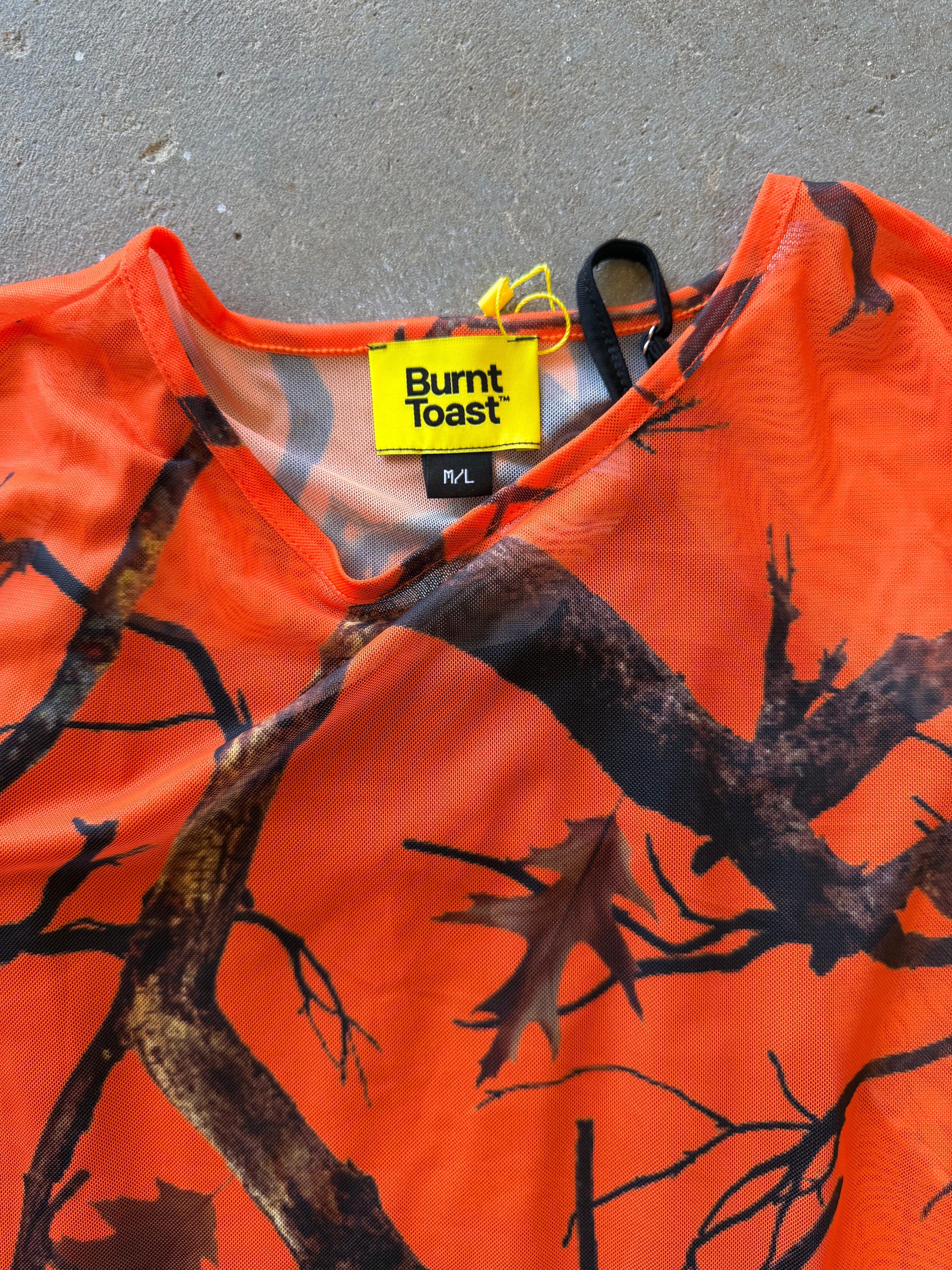 An orange short-sleeve top with a black and brown tree branch and leaf camouflage pattern, featuring a yellow Burnt Toast brand label at the collar