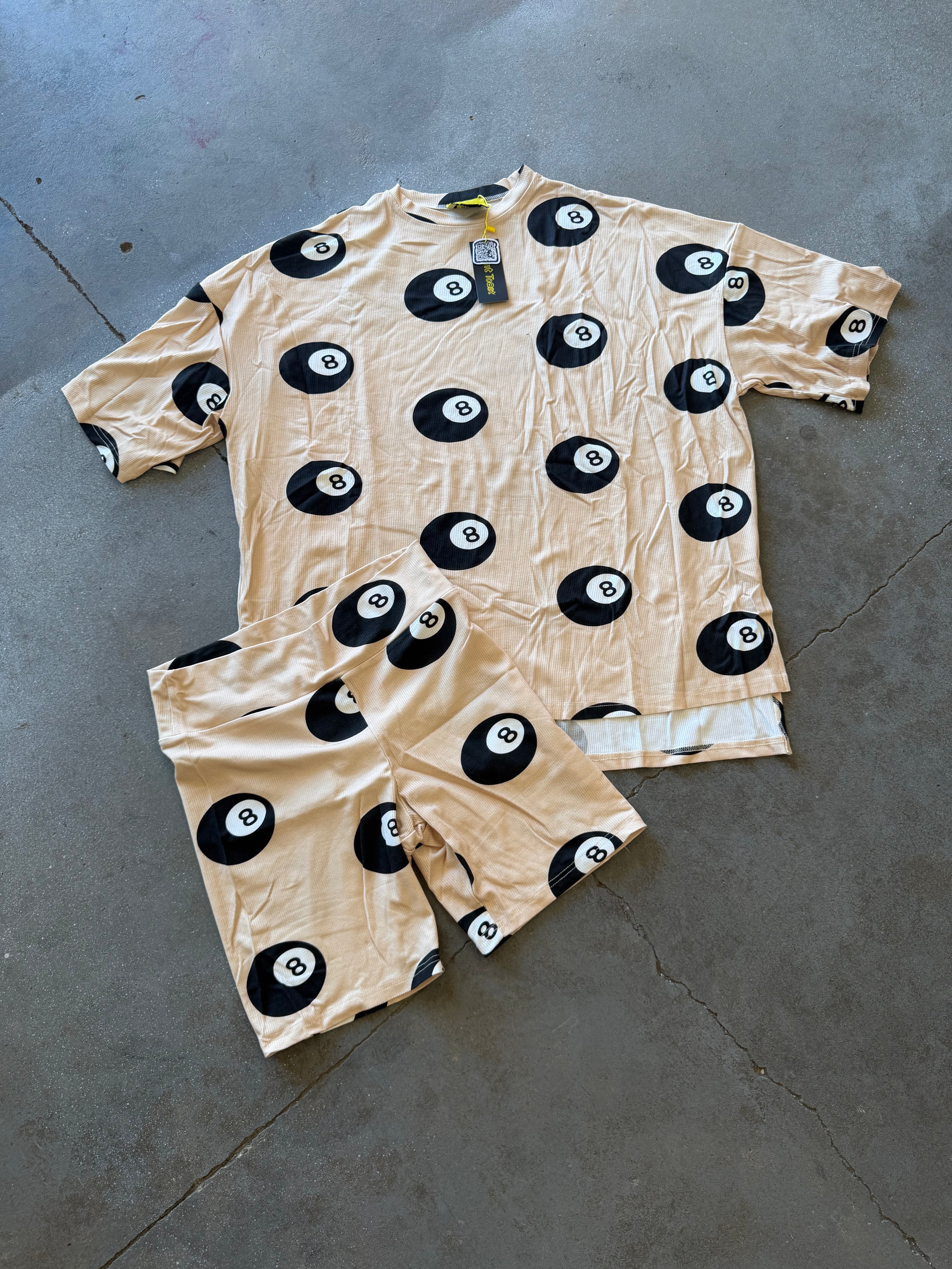 A two-piece matching pajama set laid flat on a gray concrete floor. The set includes a short-sleeve top and shorts in a light beige or peach color, both covered in a black 8-ball pattern