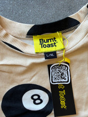 Close-up of a beige ribbed knit top with black graphic print, featuring a large billiard 8-ball motif and a yellow Burnt Toast brand tag with a QR code hangtag