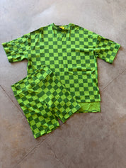 A two-piece matching set consisting of a short-sleeve t-shirt and shorts, both featuring a green and dark green checkerboard pattern, laid flat on a concrete floor