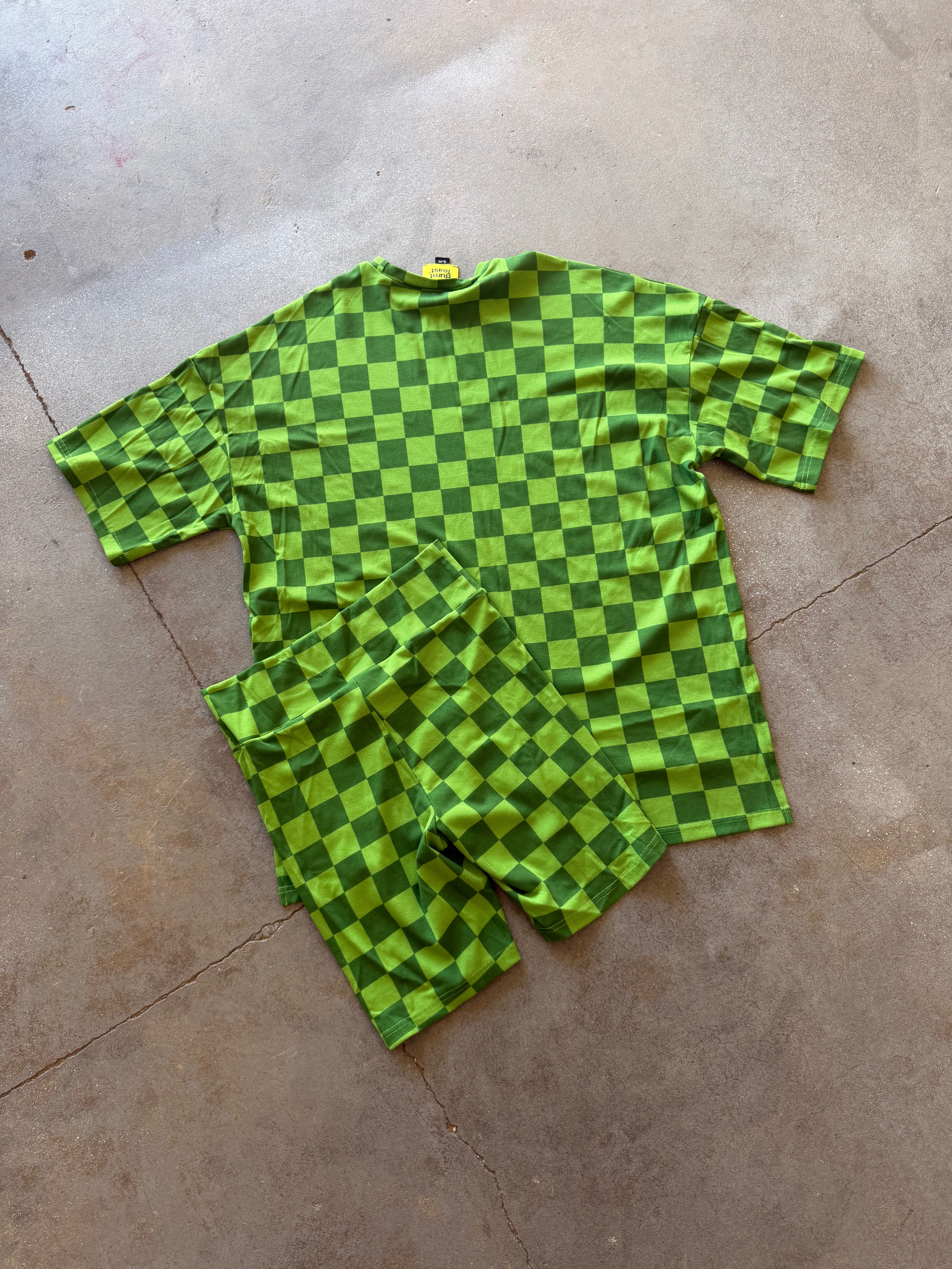 A two-piece matching set consisting of a short-sleeve t-shirt and shorts, both featuring a green and dark green checkerboard pattern, laid flat on a concrete floor