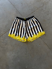 Black and white striped shorts with a bright yellow lace hem, laid flat on a gray concrete floor