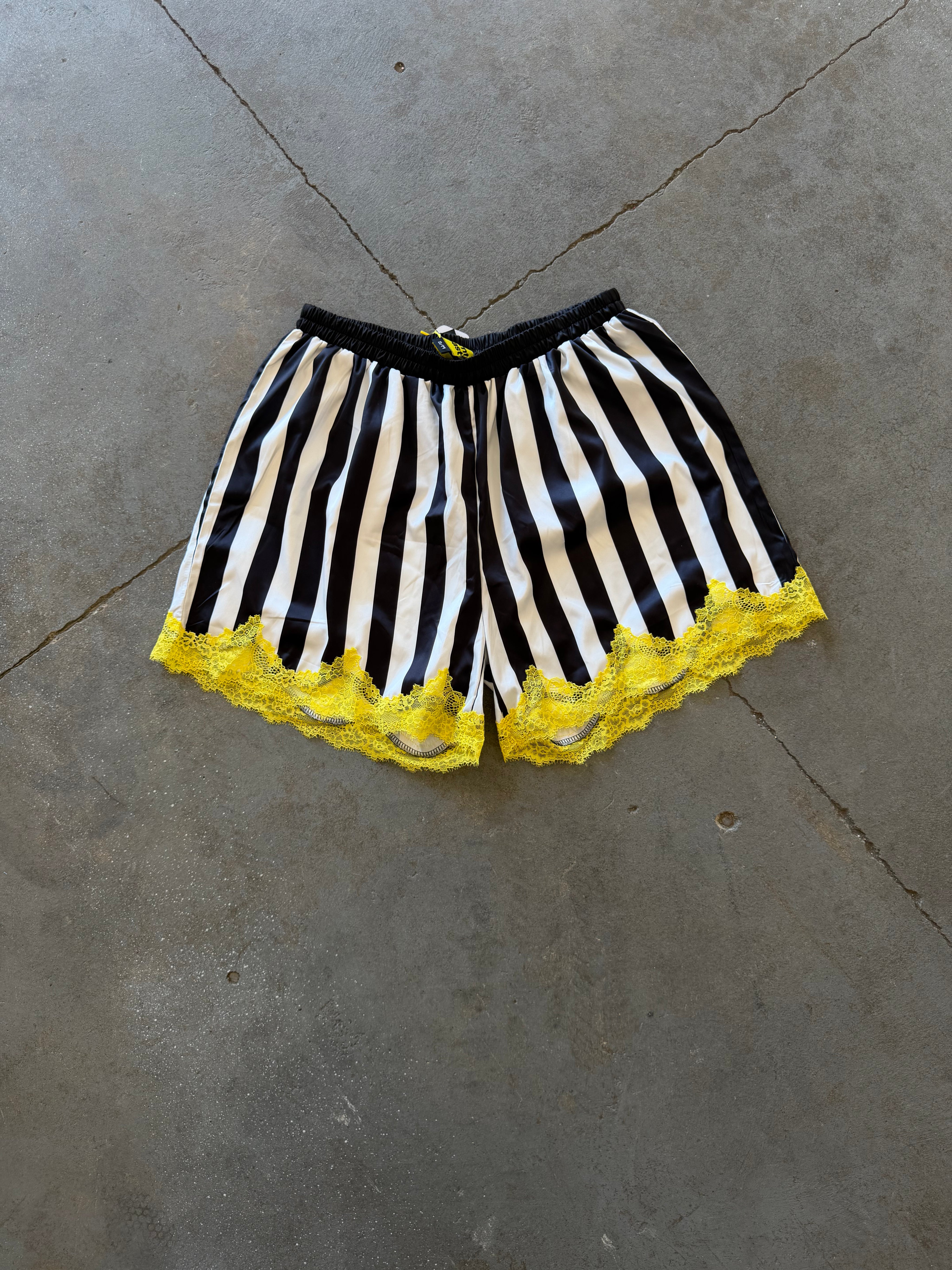 Black and white striped shorts with a bright yellow lace hem, laid flat on a gray concrete floor