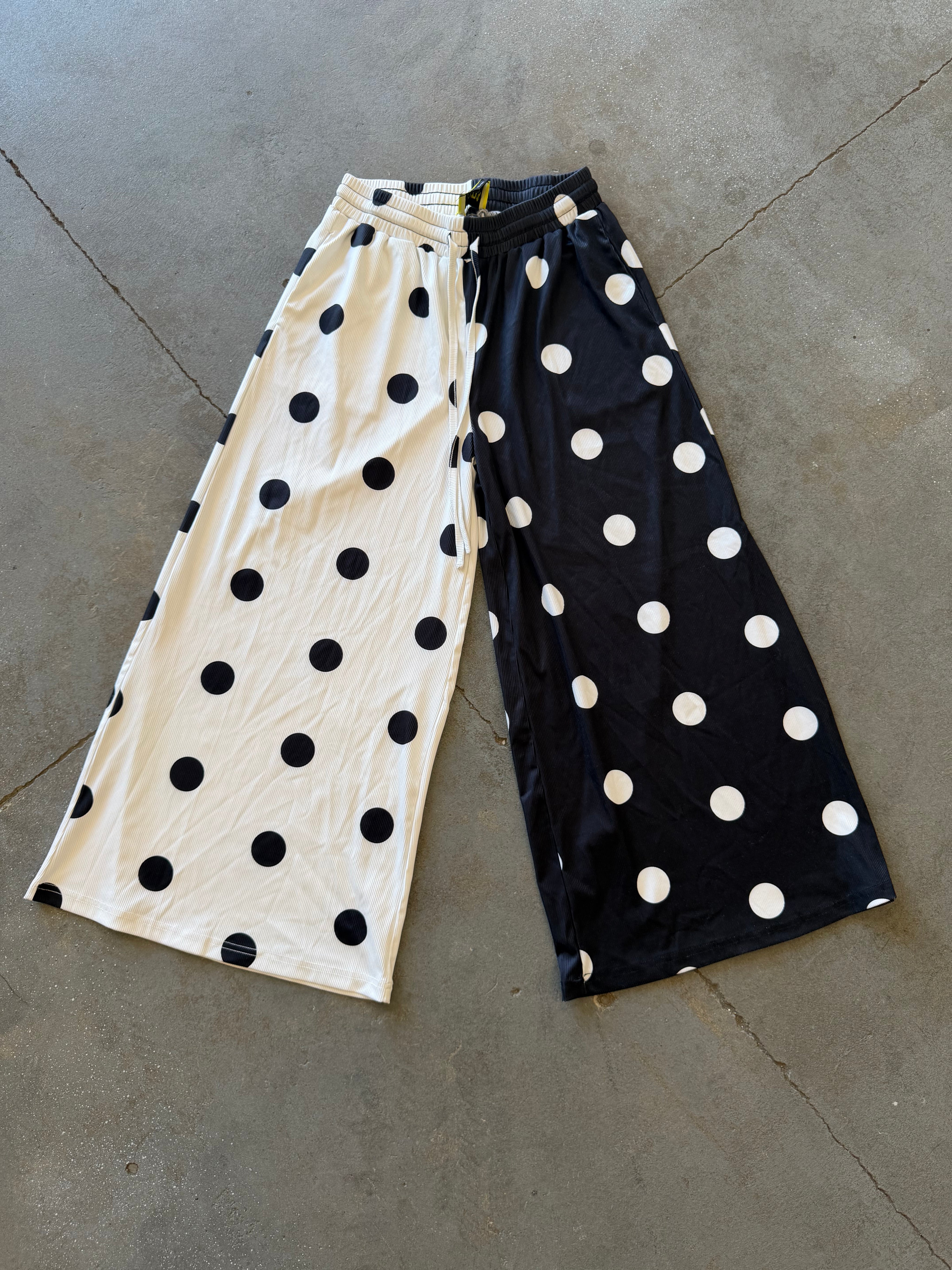 A pair of wide-leg cropped pants laid flat on a gray concrete floor, featuring a split design with one leg in cream with black polka dots and the other in black with white polka dots
